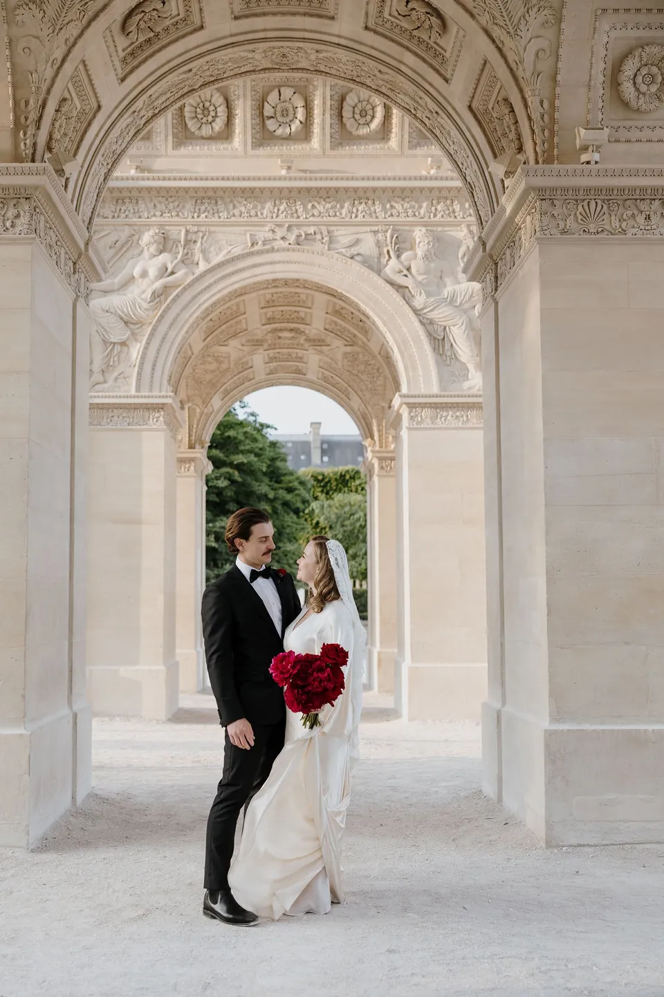 Kara and Samuel standing together beneath a carved stone archway, with her red bouquet by Ivy Bleu, at the Louvre in Paris, France, during an Elopement Collective elopement. Photograph by George Bowden.