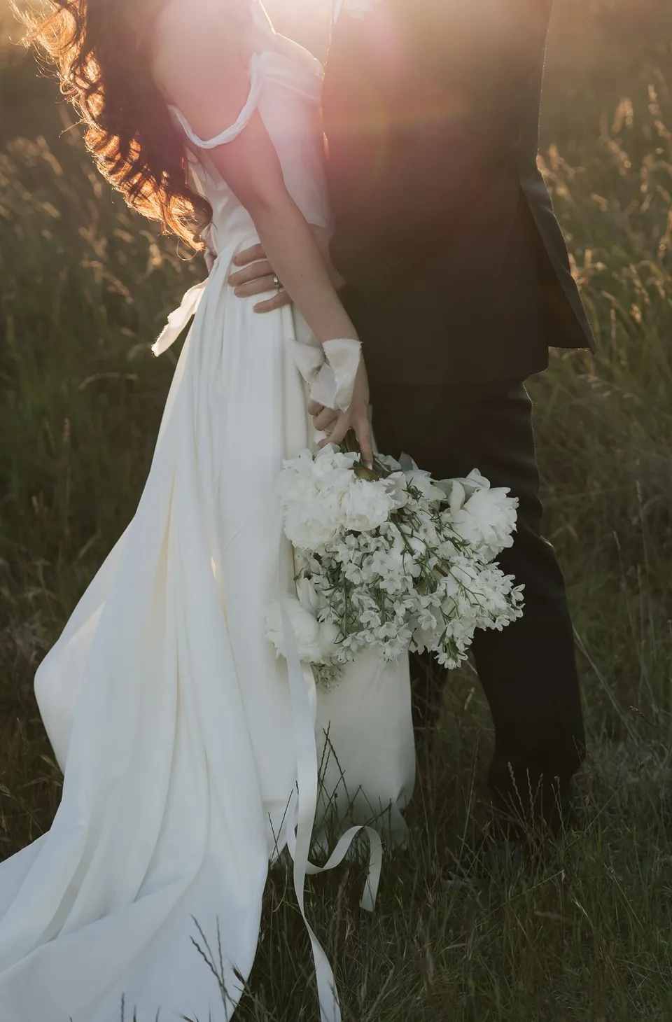 Warm sunlight on Clara’s white dress and bouquet by The Romantics as she stands beside Harry, at Thalia Haven on the East Coast of Tasmania, for an Elopement Collective elopement. Photograph by Bulb Creative.
