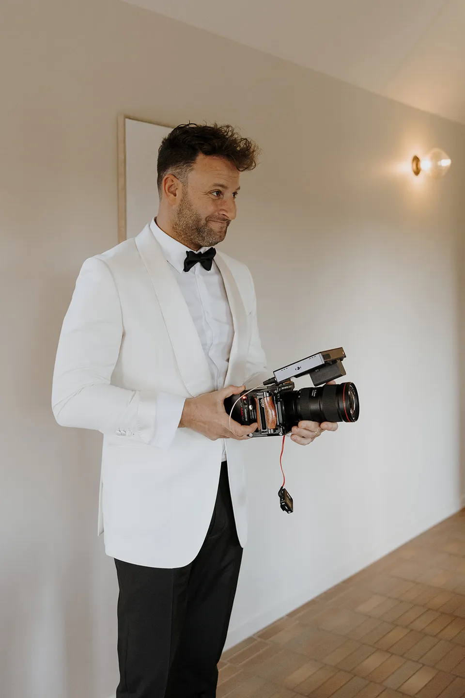 Greg holding a camera inside the cabin, wearing a white dinner jacket and black bow tie at Roundhill Retreat in Obi Obi, Sunshine Coast Hinterland, for their Elopement Collective elopement.