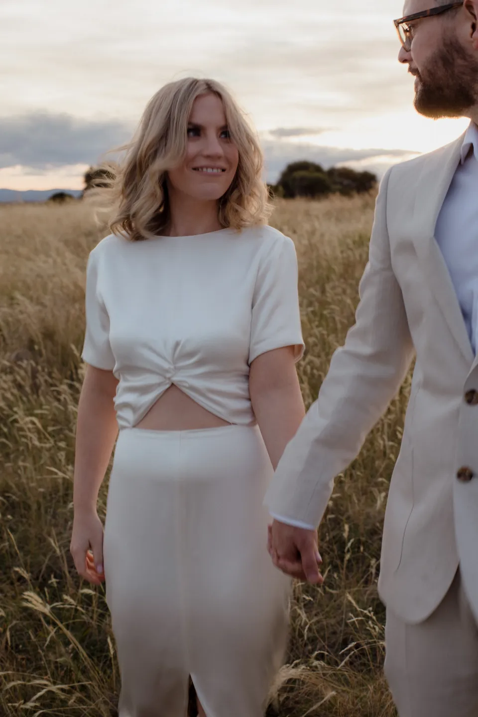 Jemmah smiling towards Jack as they hold hands among the grasses, at Thalia Haven on the East Coast of Tasmania, Australia, for an Elopement Collective elopement. Photograph by Joey Willis.