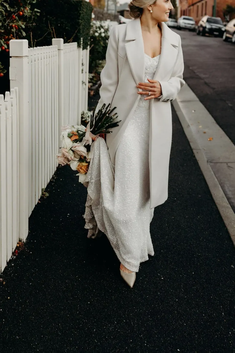 A bride in a pale coat carries a bouquet beside a white picket fence.