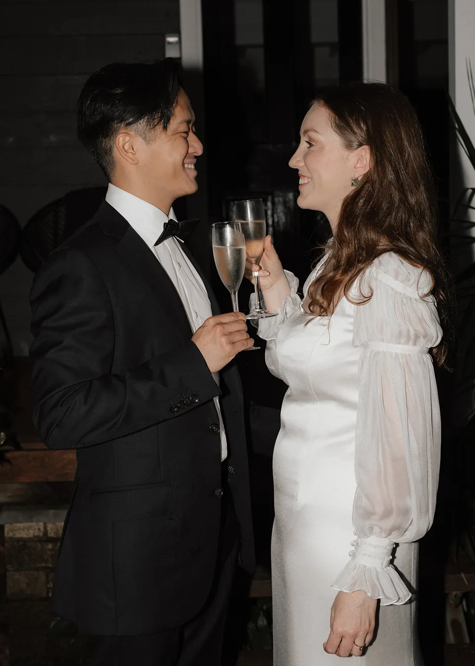 The couple smiling at each other as they toast beside the verandah. At Fig Tree Villa in Ewingsdale, Byron Bay, for an Elopement Collective elopement. Photograph by Bulb Creative.