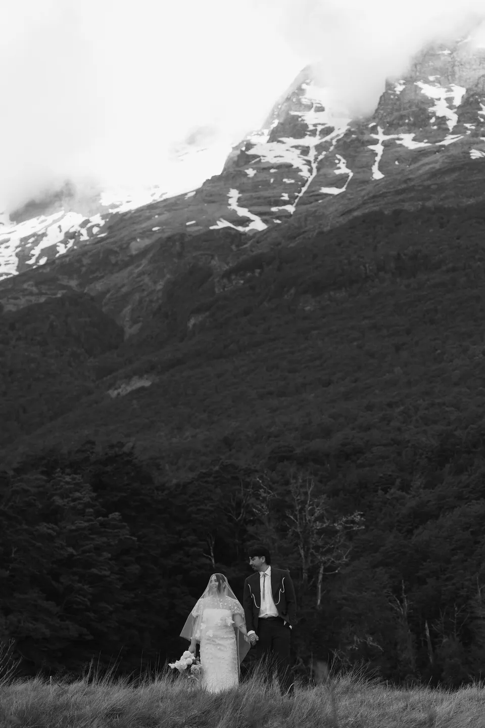 A black and white photograph of Jaeda and Adrian standing together beneath a sweep of snow-streaked mountains and forest, in the field on the road to Paradise near Glenorchy, in the Queenstown region of New Zealand, for an Elopement Collective Queenstown elopement. Photograph by Bulb Creative.