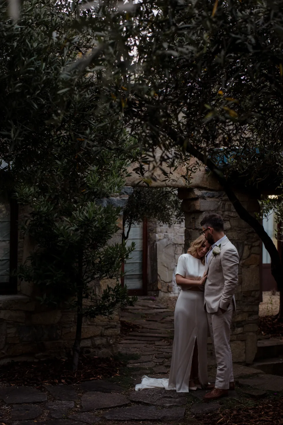 The couple standing beneath trees in the shaded stone courtyard, at Thalia Haven on the East Coast of Tasmania, Australia, for an Elopement Collective elopement. Photograph by Joey Willis.
