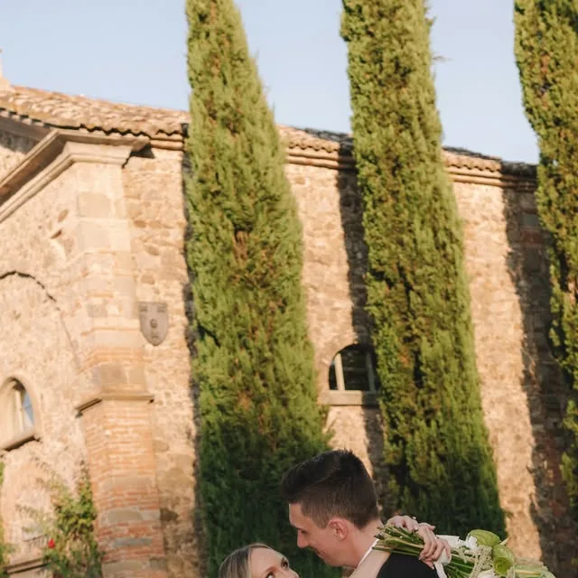 A couple embracing outside a stone building framed by tall trees.