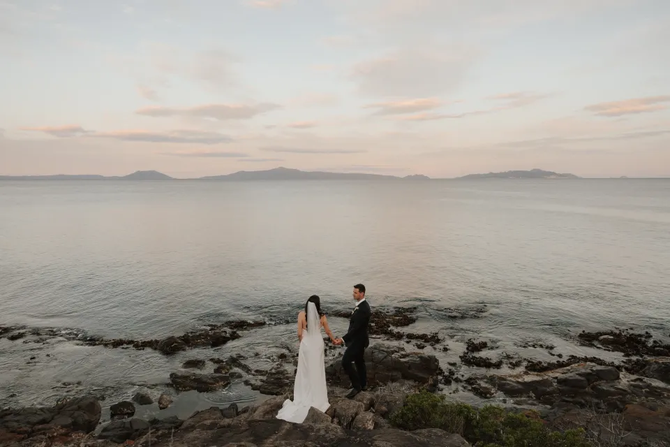 Dani and Anthony standing together above the rocky shore as soft pink clouds stretch across Great Oyster Bay, at Thalia Haven on Tasmania’s east coast, for an Elopement Collective elopement. Photograph by Bulb Creative.