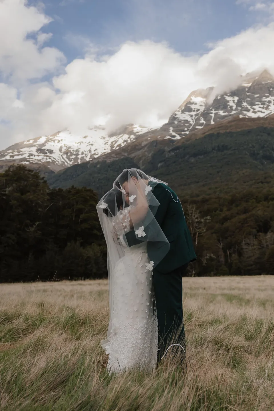 Jaeda and Adrian kissing beneath her sheer flower-appliqué veil in the golden grass, with snowy mountains behind them, in the field on the road to Paradise near Glenorchy, in the Queenstown region of New Zealand, for an Elopement Collective Queenstown elopement. Photograph by Bulb Creative.