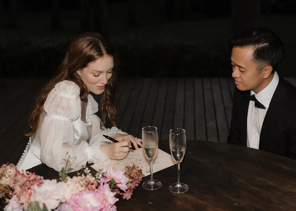 Elyse signing paperwork at a timber table with flowers and glasses beside her. At Fig Tree Villa in Ewingsdale, Byron Bay, for an Elopement Collective elopement. Photograph by Bulb Creative.