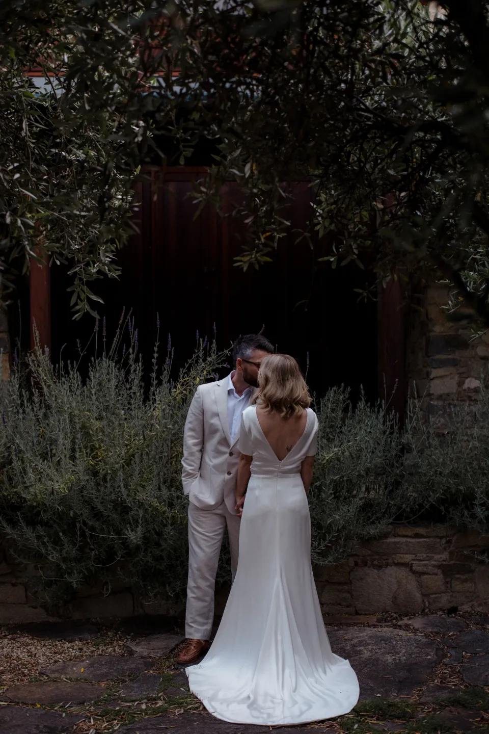 Jemmah and Jack embracing beneath leafy courtyard trees, at Thalia Haven on the East Coast of Tasmania, Australia, for an Elopement Collective elopement. Photograph by Joey Willis.