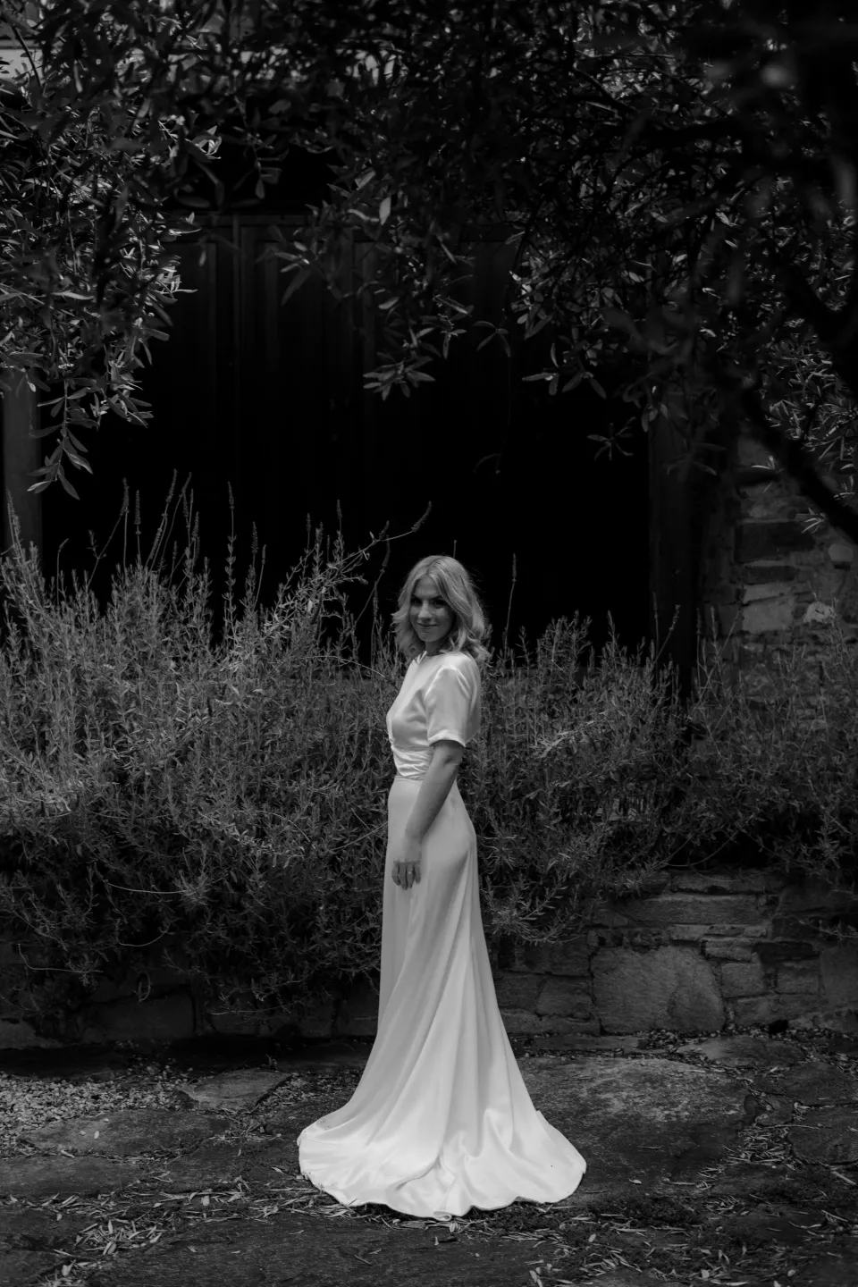 Black and white portrait of Jemmah standing in the courtyard with her dress train behind her, at Thalia Haven on the East Coast of Tasmania, Australia, for an Elopement Collective elopement. Photograph by Joey Willis.