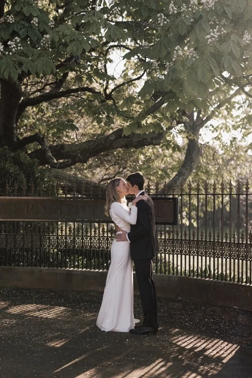 A couple kiss beneath leafy branches beside an iron garden fence.