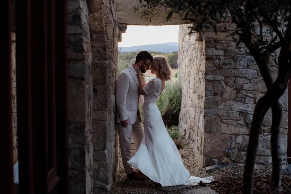 Jemmah and Jack kissing beneath a stone lintel with the meadow beyond, at Thalia Haven on the East Coast of Tasmania, Australia, for an Elopement Collective elopement. Photograph by Joey Willis.