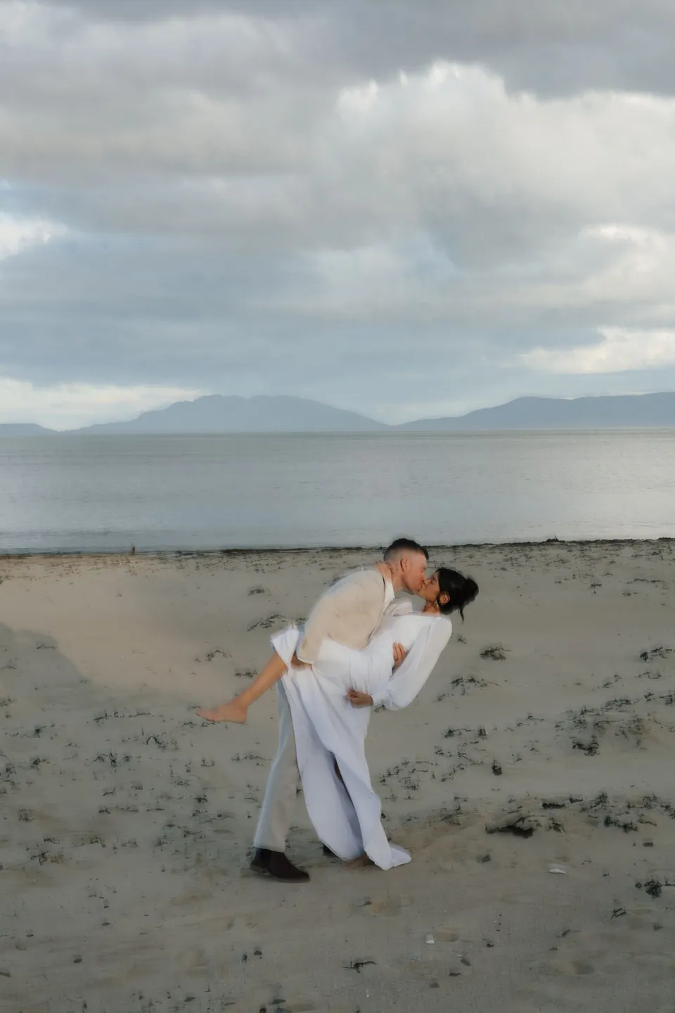 Mitch dipping Renee into a kiss on the sand, with calm water and distant hills behind them. At Piermont Retreat on Tasmania’s east coast, for an Elopement Collective elopement. Photograph by Bulb Creative.