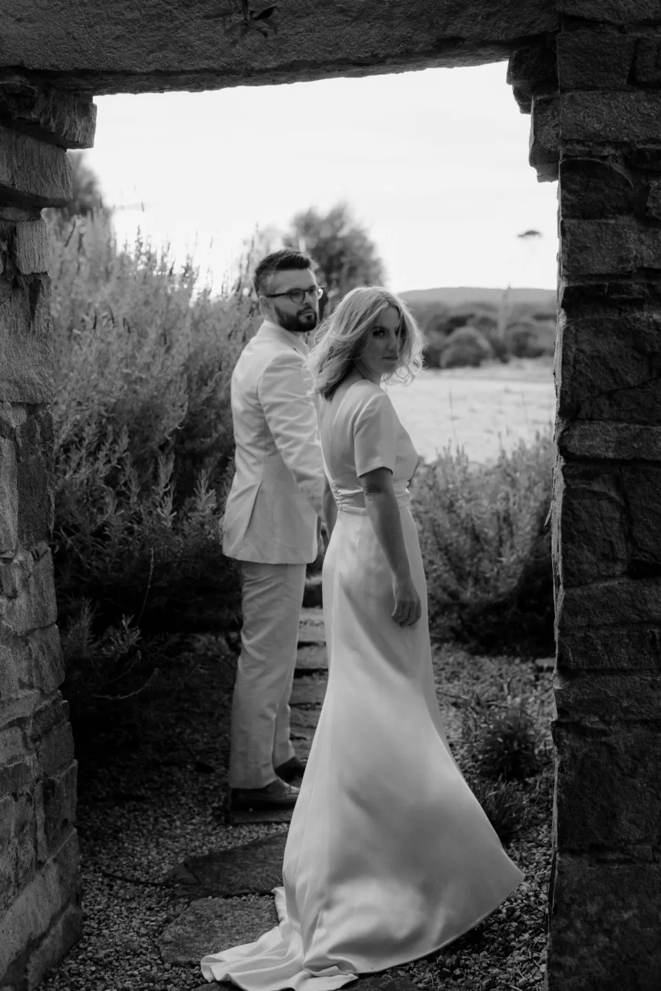 Black and white photograph of Jemmah and Jack kissing beneath a stone doorway, at Thalia Haven on the East Coast of Tasmania, Australia, for an Elopement Collective elopement. Photograph by Joey Willis.