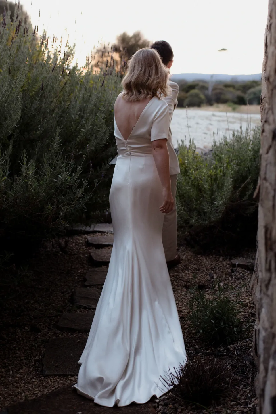 The couple standing together on a path beside tall grass, at Thalia Haven on the East Coast of Tasmania, Australia, for an Elopement Collective elopement. Photograph by Joey Willis.