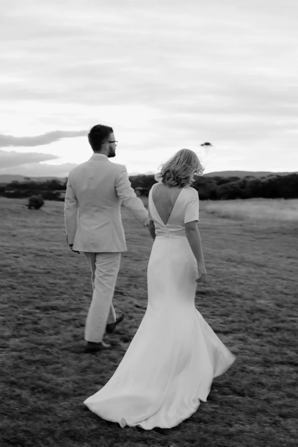 Black and white rear view of the couple walking out into the coastal meadow, at Thalia Haven on the East Coast of Tasmania, Australia, for an Elopement Collective elopement. Photograph by Joey Willis.
