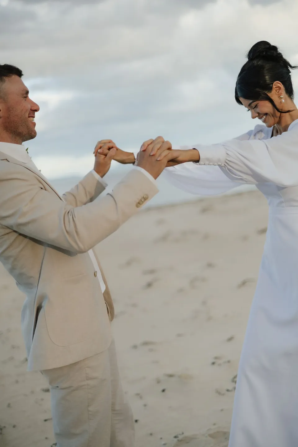Renee and Mitch holding both hands and laughing together on the beach. At Piermont Retreat on Tasmania’s east coast, for an Elopement Collective elopement. Photograph by Bulb Creative.