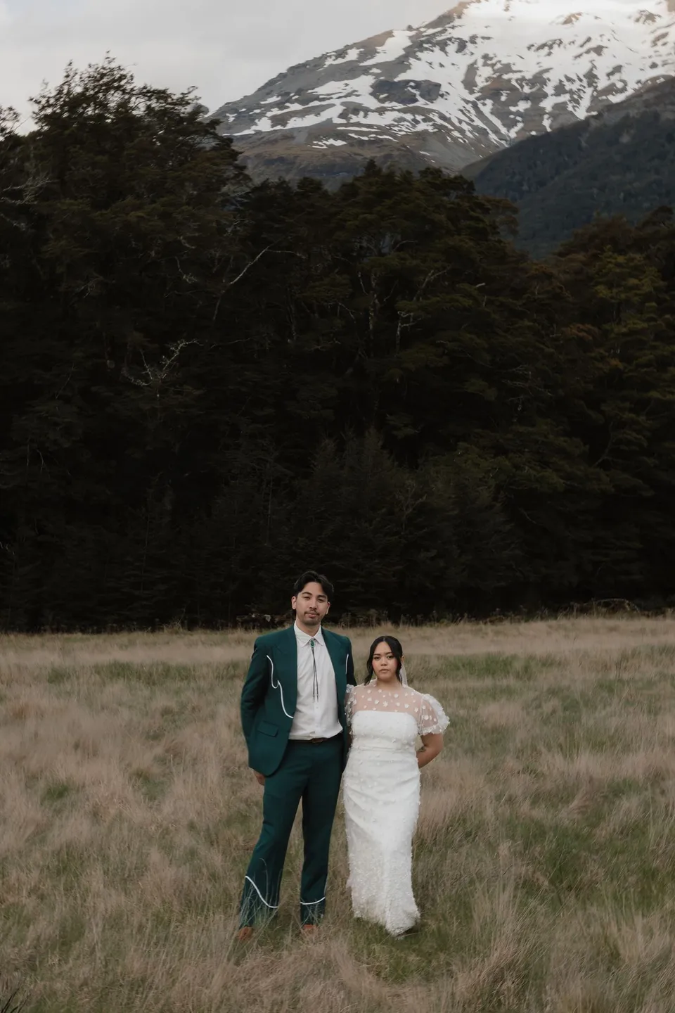 Jaeda and Adrian standing close together in the meadow, framed by forest and cloud-wrapped mountain peaks, in the field on the road to Paradise near Glenorchy, in the Queenstown region of New Zealand, for an Elopement Collective Queenstown elopement. Photograph by Bulb Creative.
