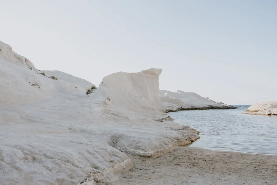 Pale sculpted rock formations surrounding a narrow inlet of calm blue water, at Sarakiniko on Milos in the Cyclades, Greece, during an Elopement Collective elopement. Photograph by George Bowden.