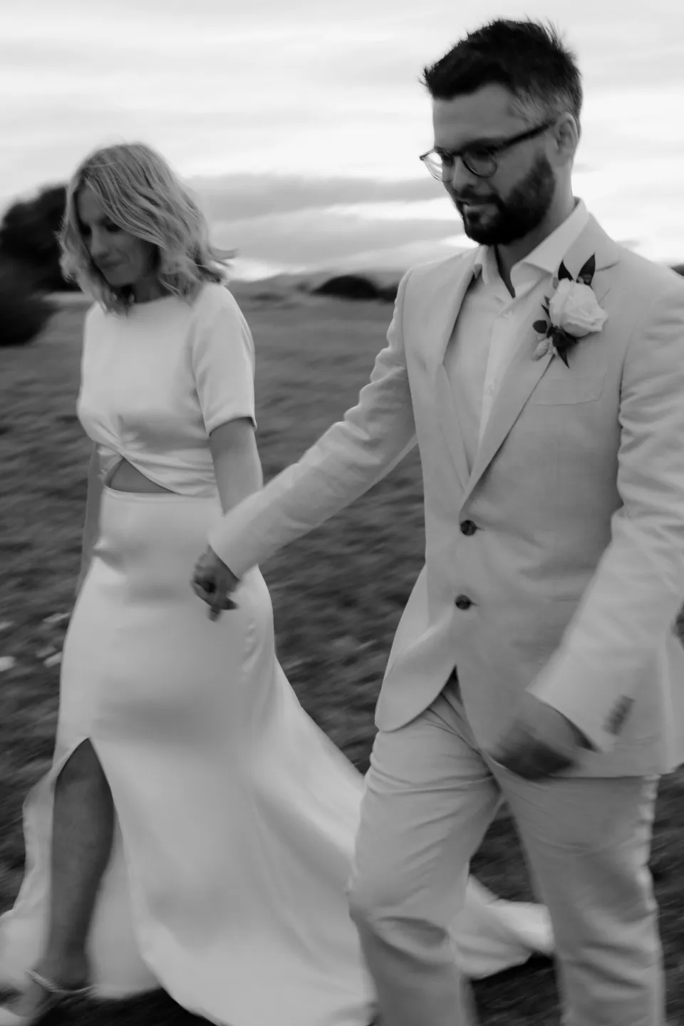 Black and white photograph of Jemmah and Jack walking hand in hand, at Thalia Haven on the East Coast of Tasmania, Australia, for an Elopement Collective elopement. Photograph by Joey Willis.