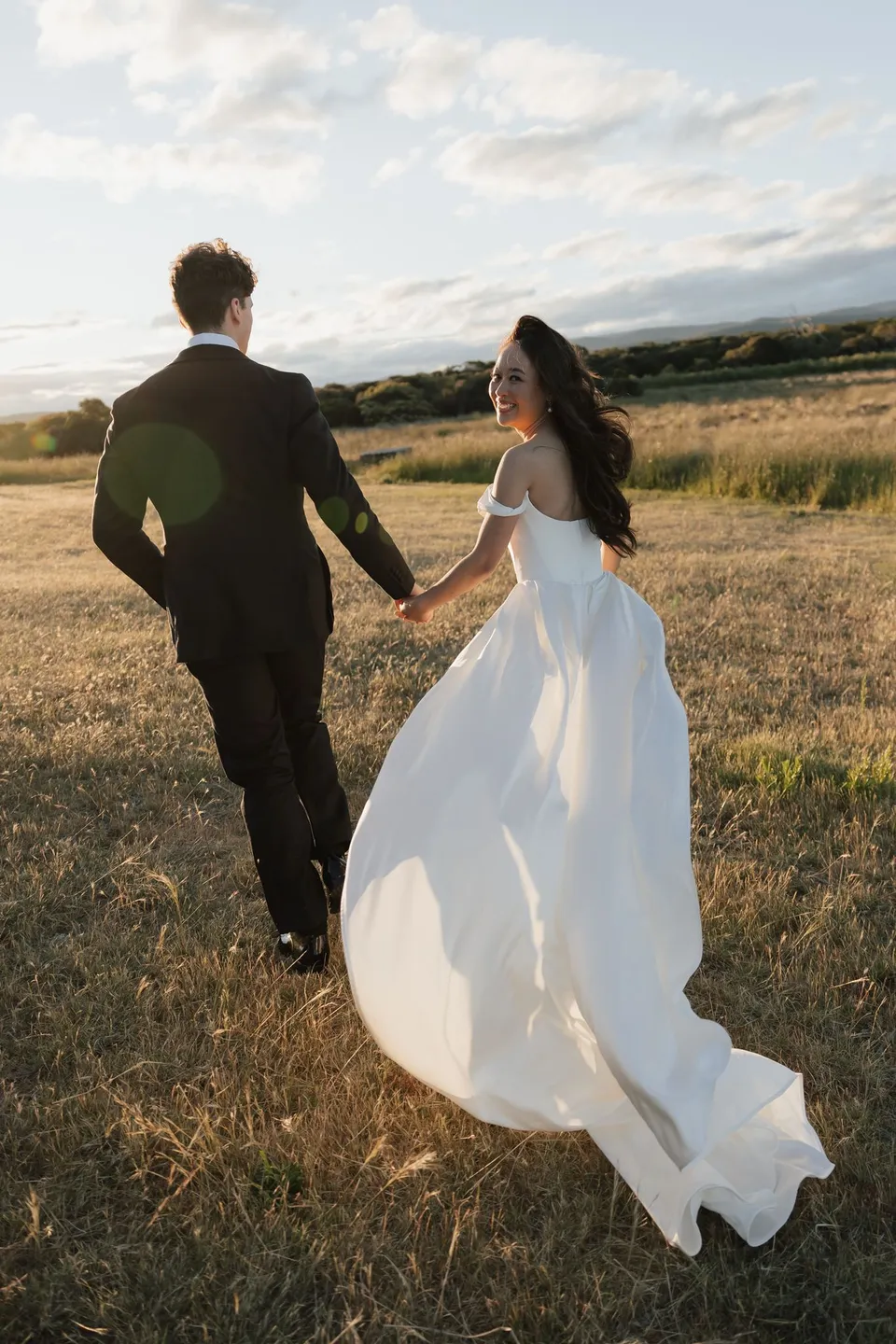 Rear view of Clara and Harry walking hand in hand through golden grass, with her dress lifted by the breeze, at Thalia Haven on the East Coast of Tasmania, for an Elopement Collective elopement. Photograph by Bulb Creative.