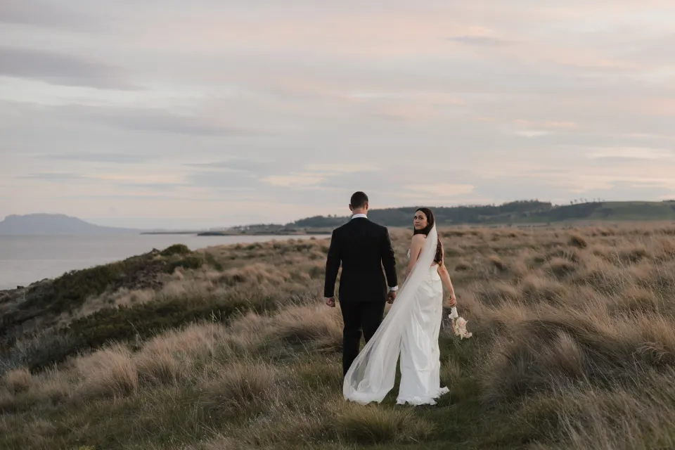 Dani looking back as she and Anthony walk hand in hand through coastal grassland beneath a pink evening sky, at Thalia Haven on Tasmania’s east coast, for an Elopement Collective elopement. Photograph by Bulb Creative.