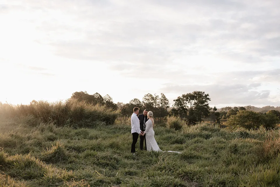 A wide view of Matt and Laura facing each other for their outdoor ceremony with celebrant Josh Withers among green grasses, in the Tweed Valley, NSW, for an Elopement Collective elopement. Photo by Bulb Creative.