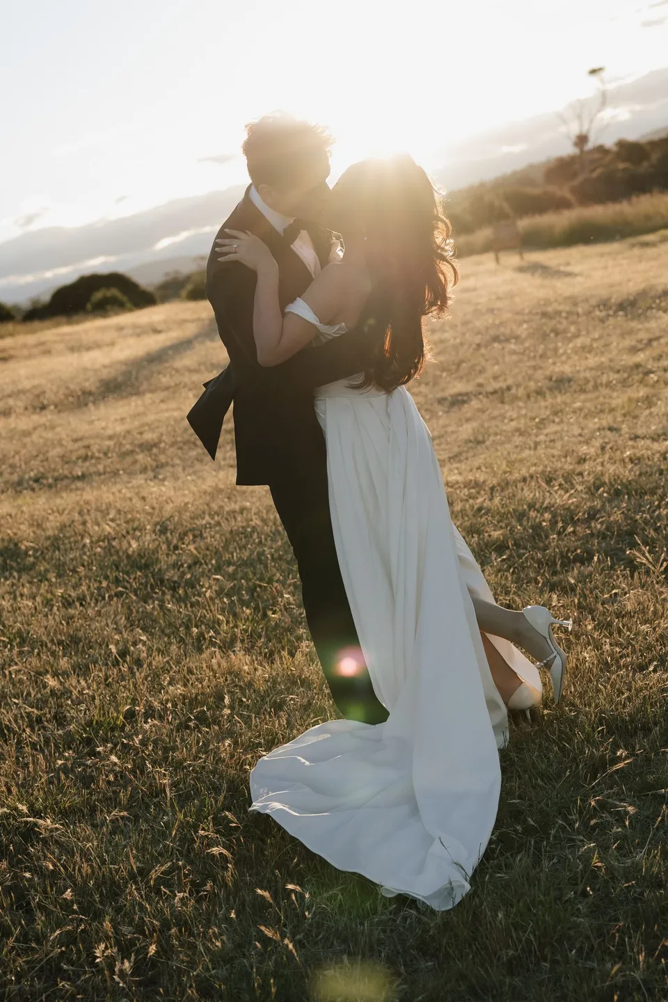 Clara and Harry kissing in golden grassland with low sunlight glowing behind them, at Thalia Haven on the East Coast of Tasmania, for an Elopement Collective elopement. Photograph by Bulb Creative.