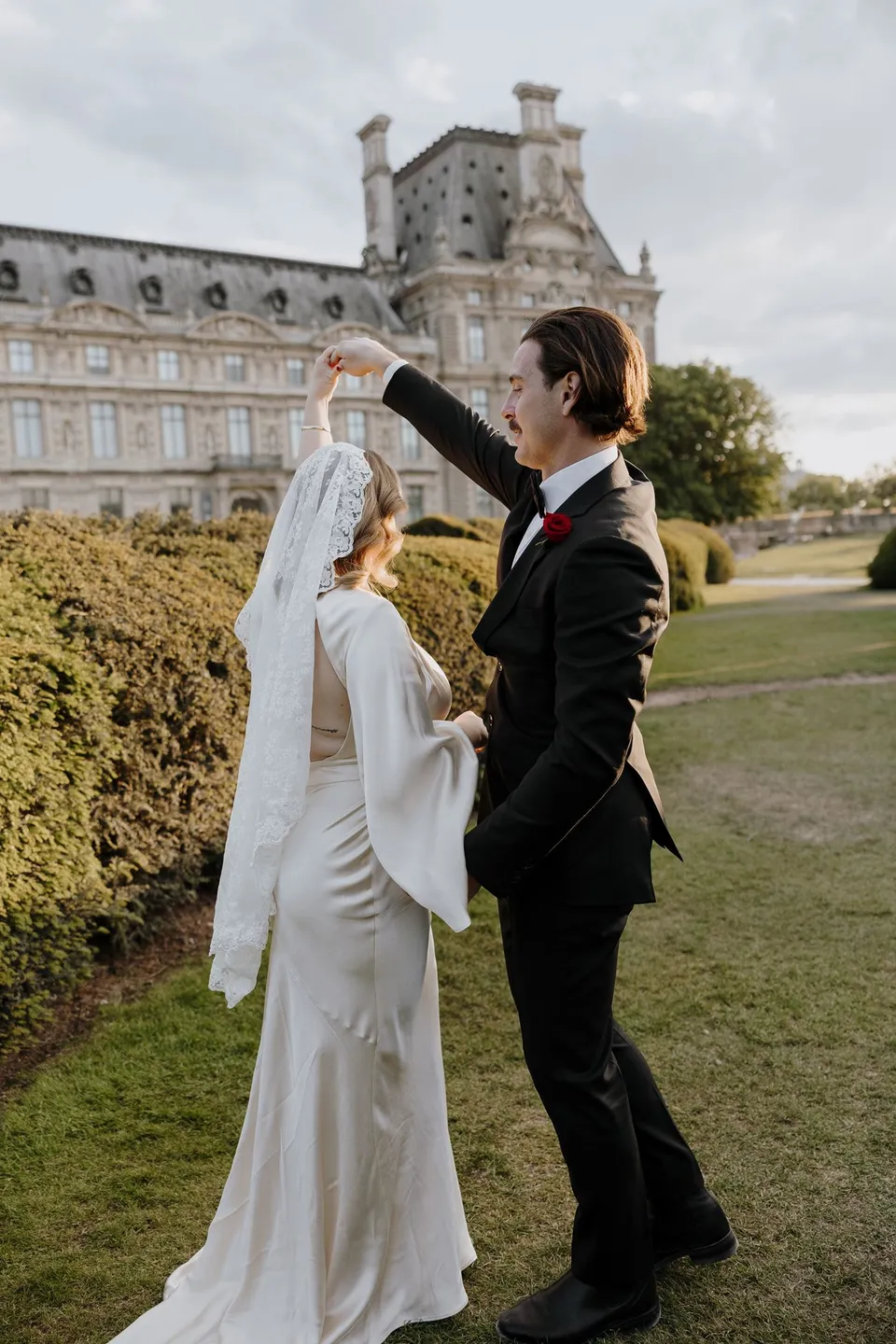 Samuel raising Kara’s hand to twirl her beside a hedge, with the Louvre’s stone facade behind them, at the Louvre in Paris, France, during an Elopement Collective elopement. Photograph by George Bowden.