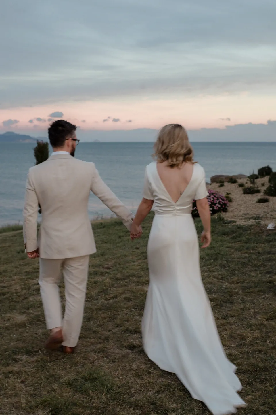 The couple walking beside the bay beneath a soft pink evening sky, at Thalia Haven on the East Coast of Tasmania, Australia, for an Elopement Collective elopement. Photograph by Joey Willis.