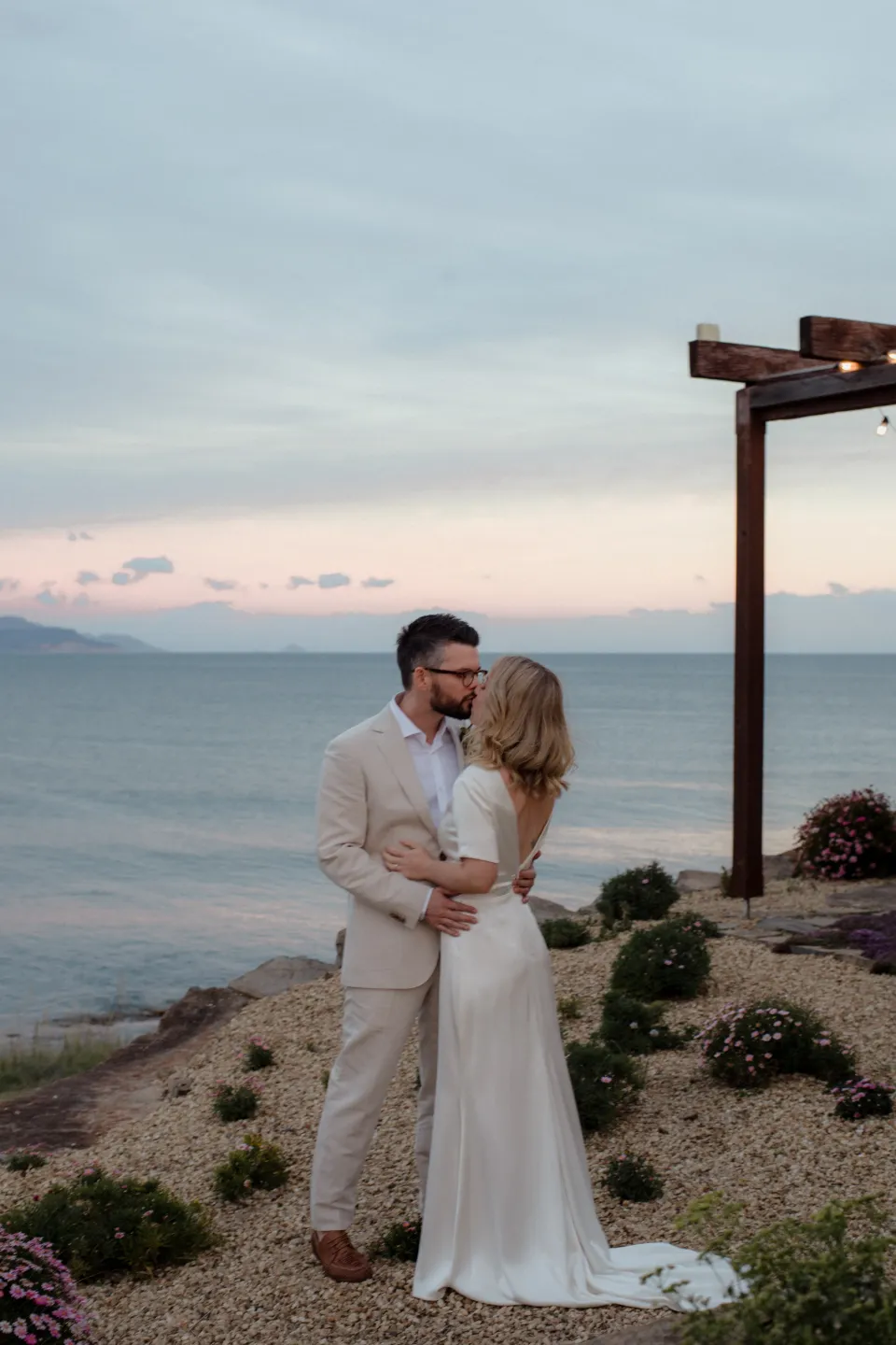 Jack holding Jemmah on the stone terrace as the sky turns pink, at Thalia Haven on the East Coast of Tasmania, Australia, for an Elopement Collective elopement. Photograph by Joey Willis.