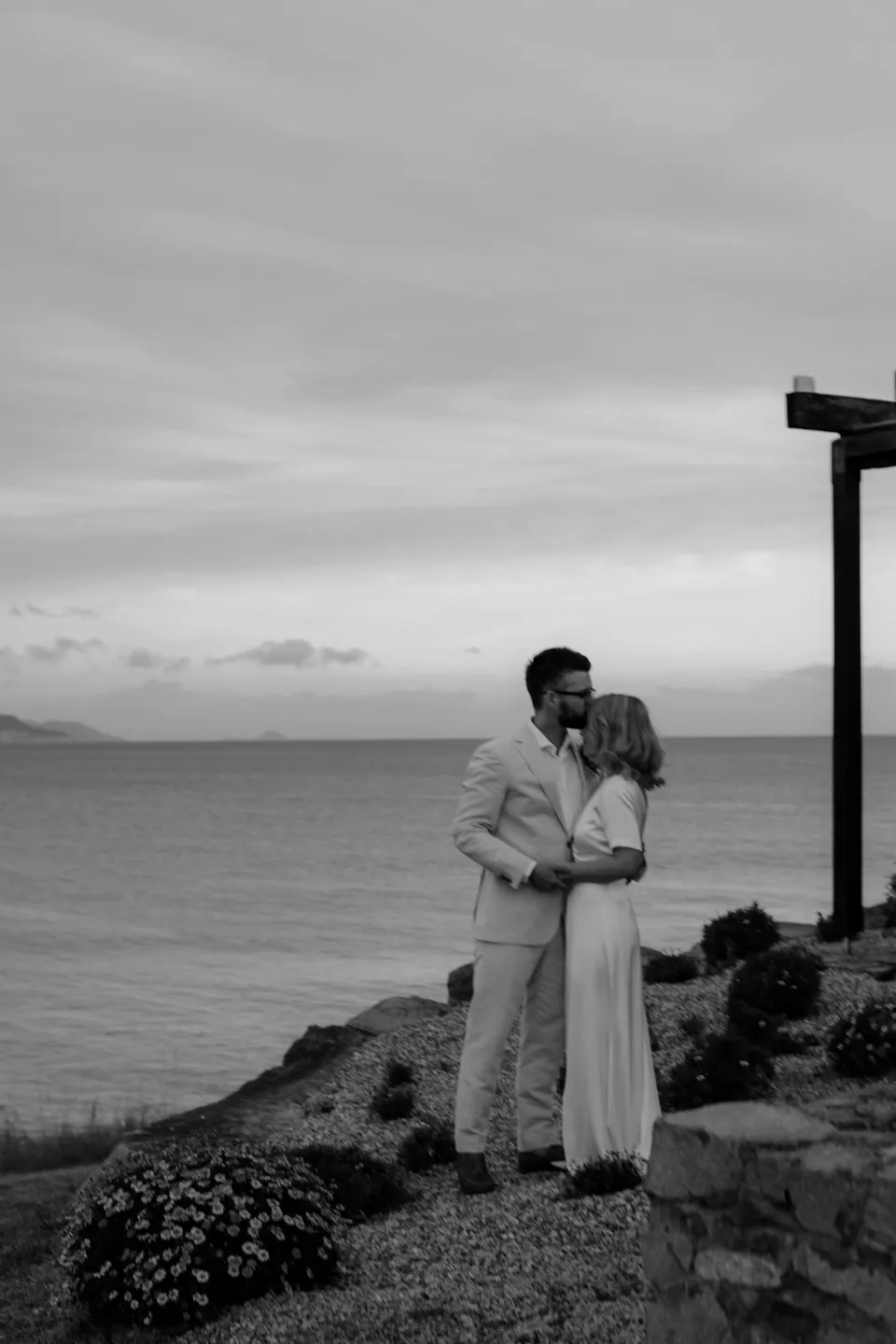 Black and white photograph of the couple embracing above the water, at Thalia Haven on the East Coast of Tasmania, Australia, for an Elopement Collective elopement. Photograph by Joey Willis.