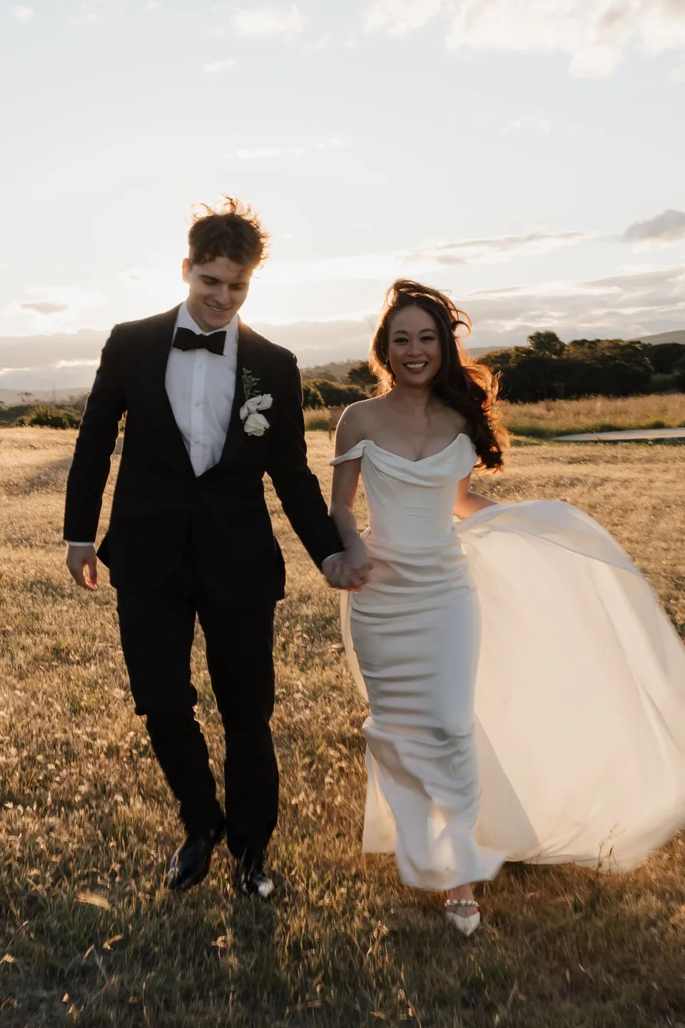 Clara and Harry smiling as they walk through the grass, with her white dress gathered in one hand, at Thalia Haven on the East Coast of Tasmania, for an Elopement Collective elopement. Photograph by Bulb Creative.