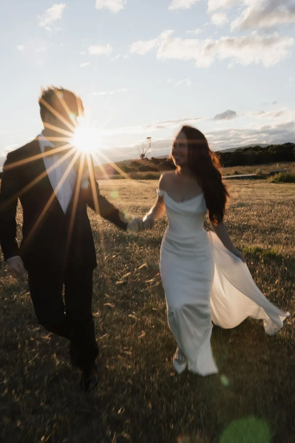 Clara and Harry walking together through golden grassland, with a sunburst beside Harry’s shoulder, at Thalia Haven on the East Coast of Tasmania, for an Elopement Collective elopement. Photograph by Bulb Creative.