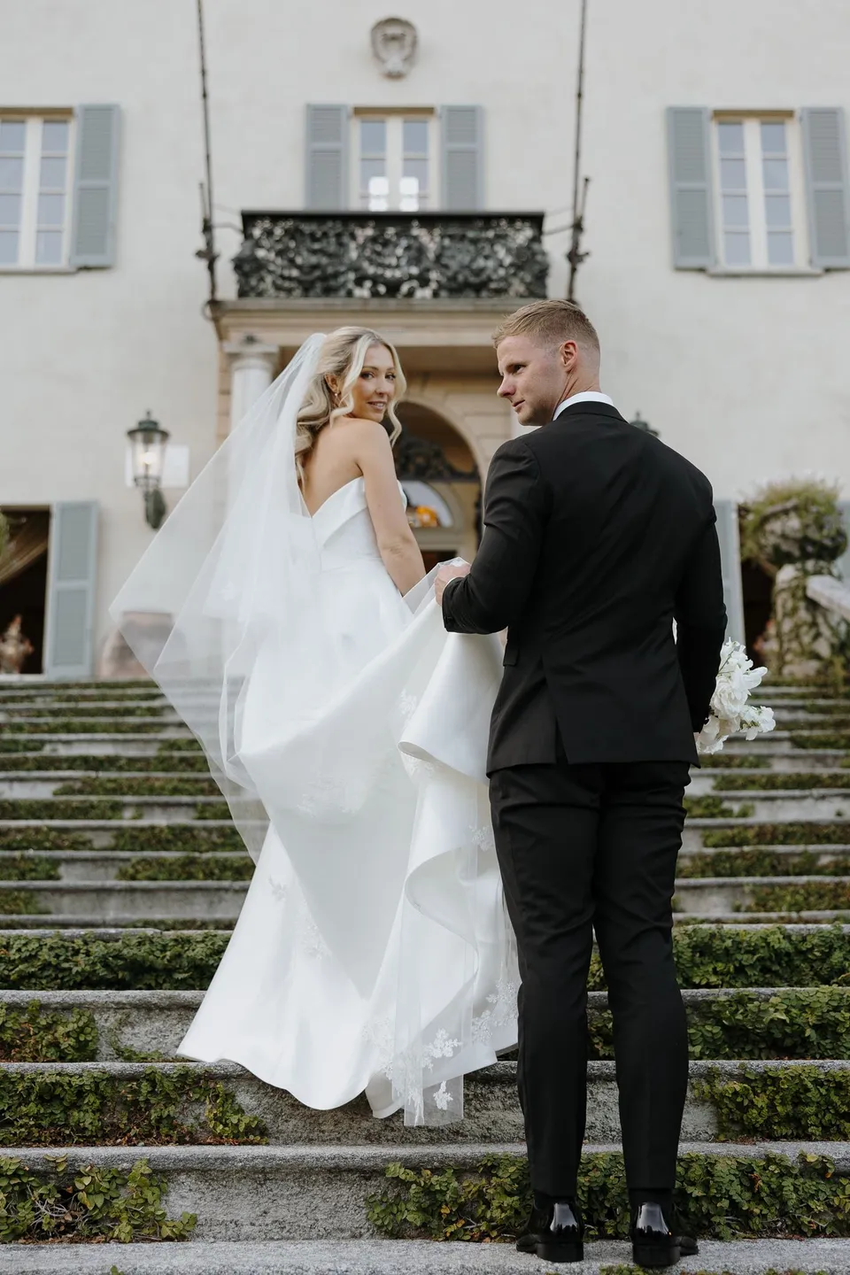 Charlton holding Zoe’s hand as they climb stone steps towards the villa, with her white veil lifting behind her, at Passalacqua in Moltrasio, Lake Como, Italy, for an Elopement Collective elopement. Photo by George Bowden.
