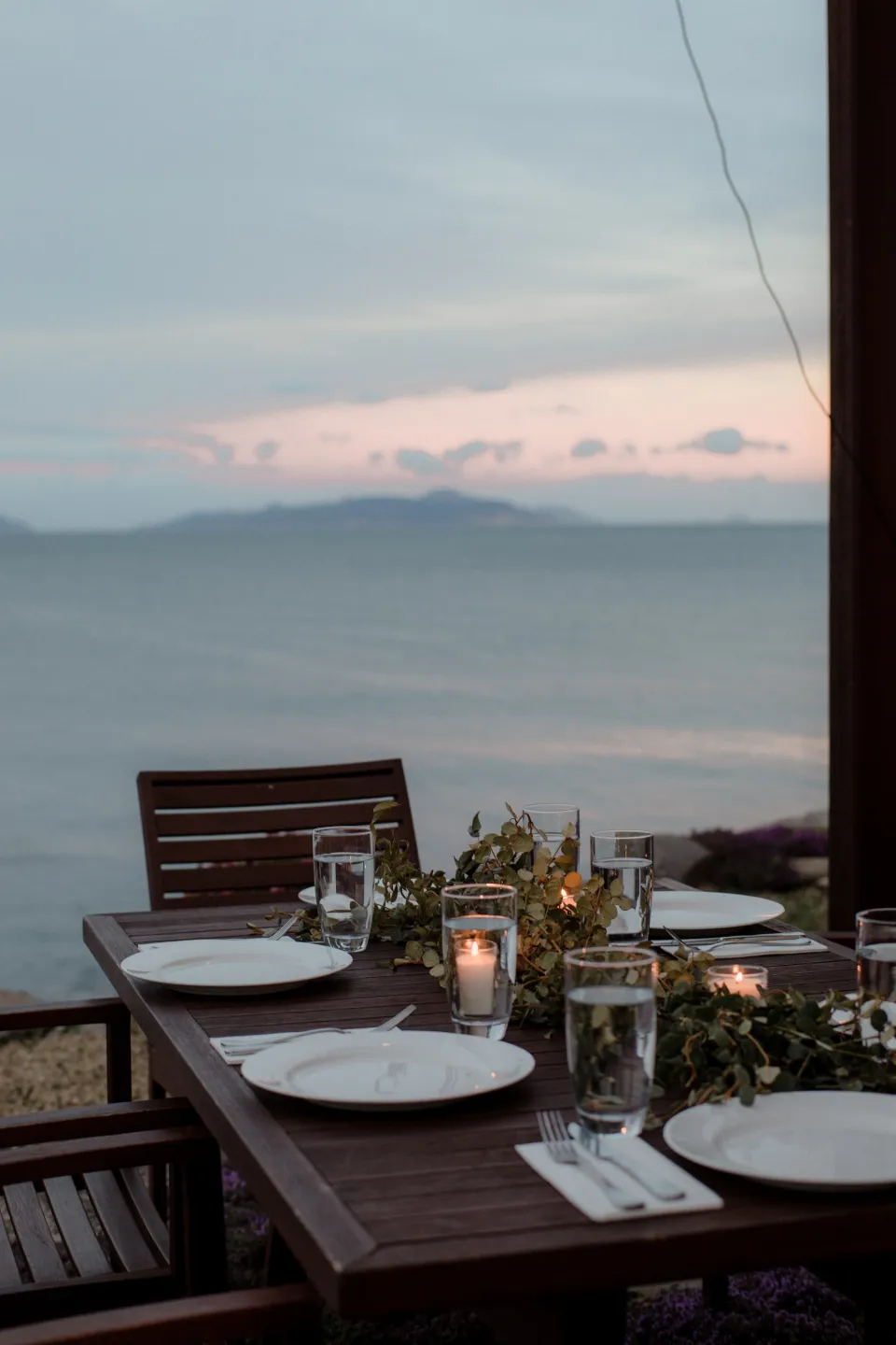 A dinner table set for the evening with the bay and pink sky beyond, at Thalia Haven on the East Coast of Tasmania, Australia, for an Elopement Collective elopement. Photograph by Joey Willis.
