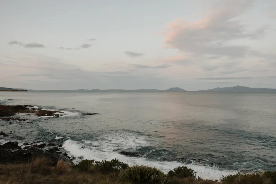Soft evening clouds above Great Oyster Bay, with foamy waves, a rocky cove and distant hills, at Thalia Haven on Tasmania’s east coast, for an Elopement Collective elopement. Photograph by Bulb Creative.