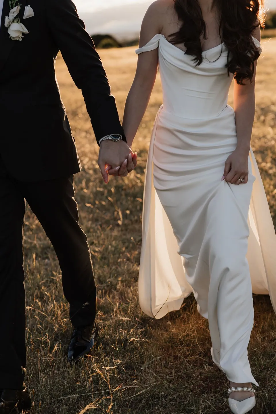 Close view of Clara and Harry holding hands as they walk, with her draped white dress, sparkling shoes and his black tuxedo, at Thalia Haven on the East Coast of Tasmania, for an Elopement Collective elopement. Photograph by Bulb Creative.