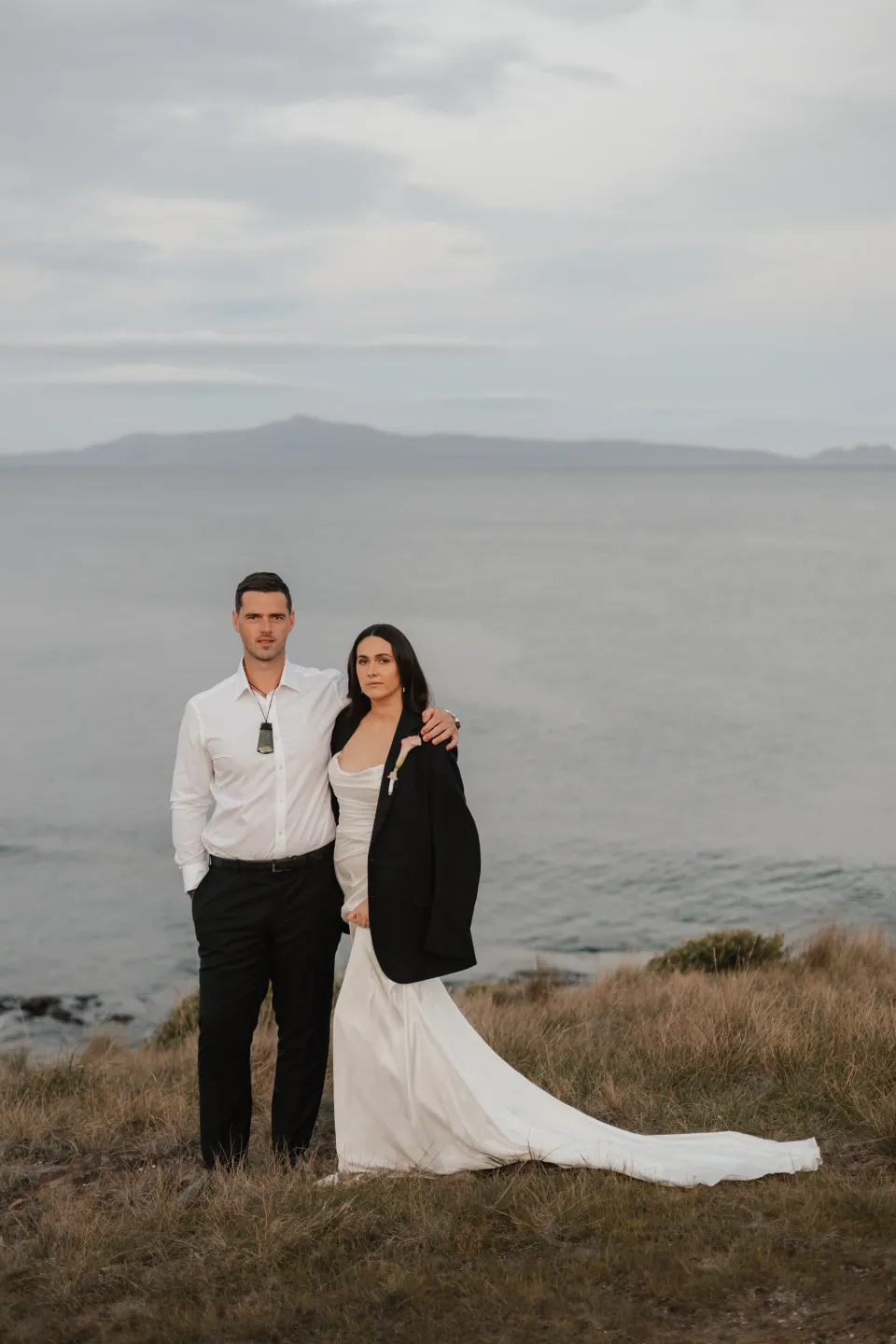 Dani wearing Anthony’s dark jacket over her wedding dress as they stand together above the bay in the evening, at Thalia Haven on Tasmania’s east coast, for an Elopement Collective elopement. Photograph by Bulb Creative.