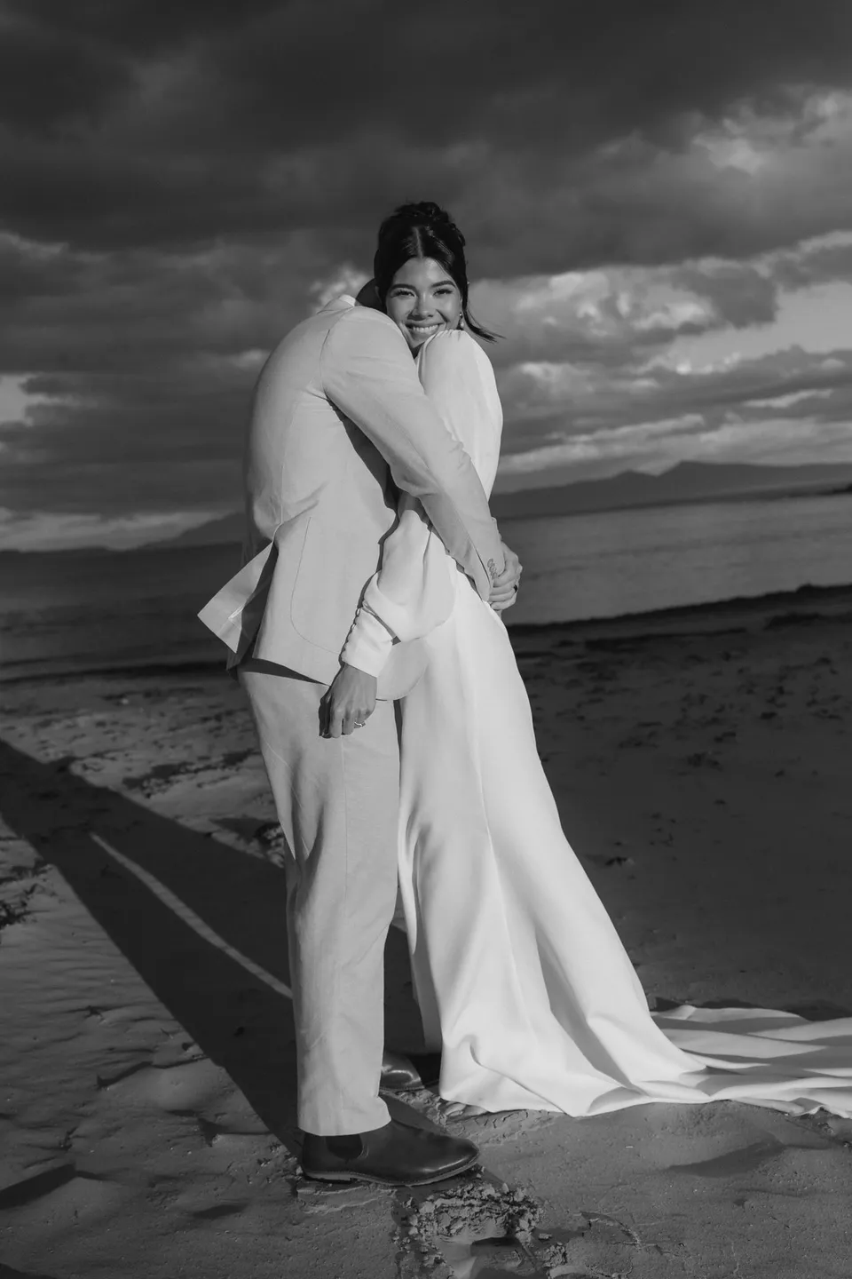 Black and white photograph of Renee laughing as Mitch leans into their embrace on the beach. At Piermont Retreat on Tasmania’s east coast, for an Elopement Collective elopement. Photograph by Bulb Creative.