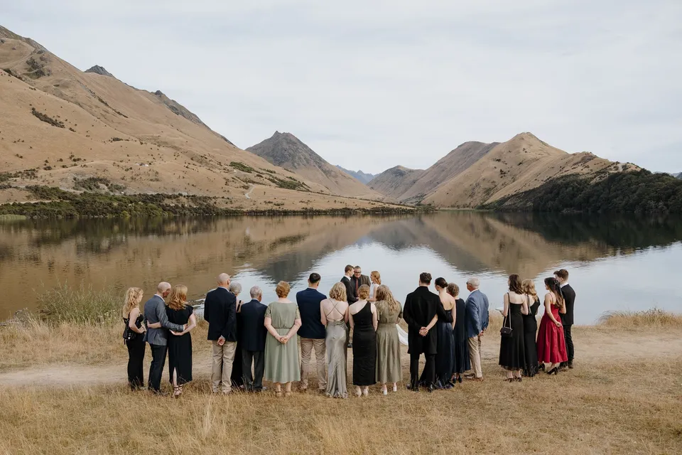 Guests gathered beside the still lake for Neve and Hamish’s ceremony, with golden mountain slopes reflected in the water, at Moke Lake in Queenstown, New Zealand, for an Elopement Collective Queenstown elopement. Photo by George Bowden.
