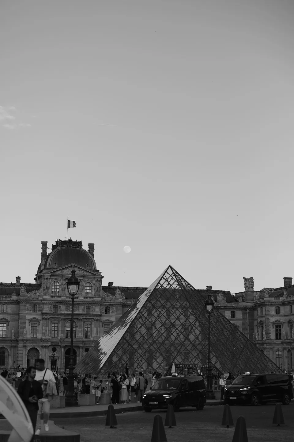 Black and white view of the Louvre’s glass pyramid, palace facade and visitors in the courtyard, at the Louvre in Paris, France, during an Elopement Collective elopement. Photograph by George Bowden.