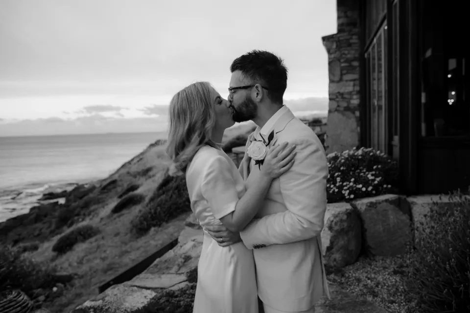 Black and white close view of Jemmah kissing Jack beneath the timber pergola, at Thalia Haven on the East Coast of Tasmania, Australia, for an Elopement Collective elopement. Photograph by Joey Willis.