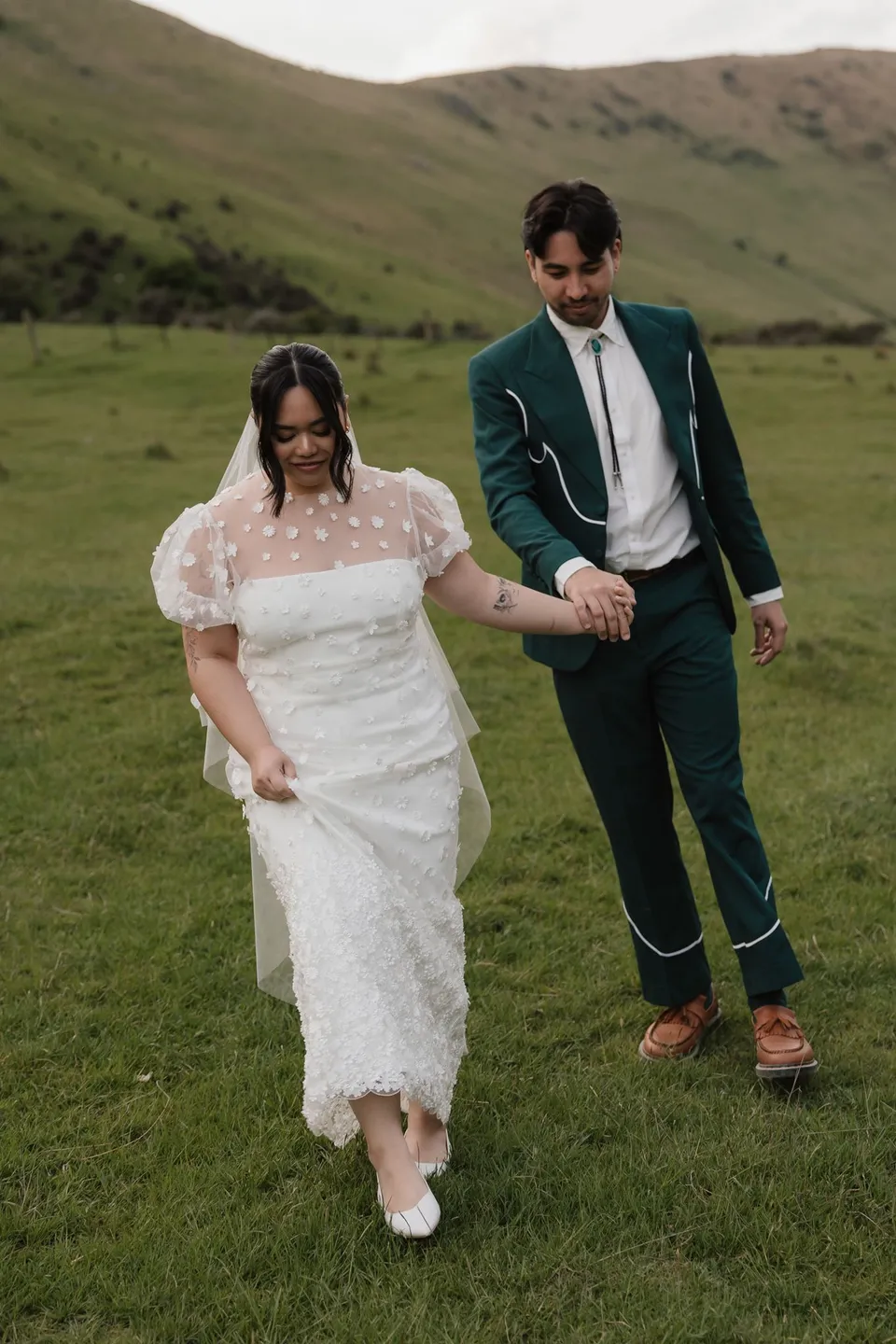 Jaeda and Adrian walking hand in hand through short green grass, with Jaeda carrying her Orange Blossom Designs bouquet, at Moke Lake, Queenstown, New Zealand, for an Elopement Collective Queenstown elopement. Photograph by Bulb Creative.