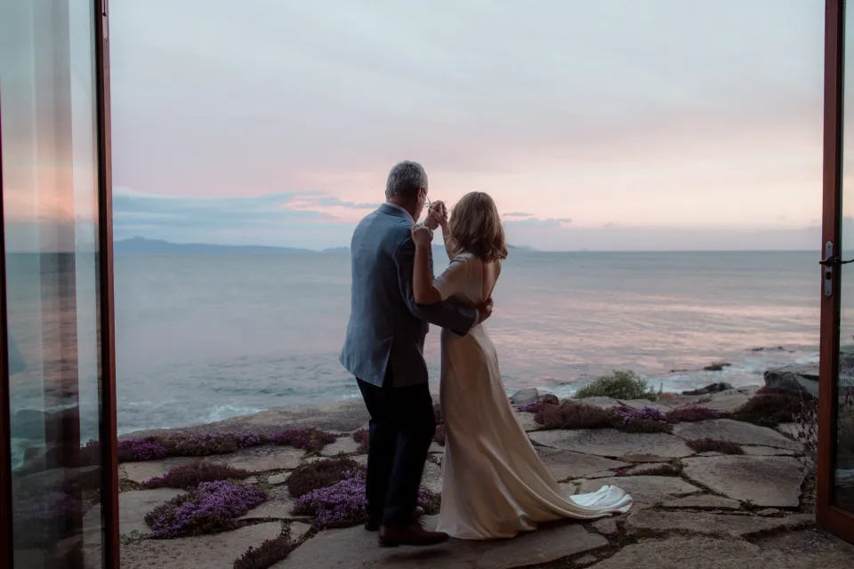 Jemmah dancing with a loved one beside the bay as seen through the open doors, at Thalia Haven on the East Coast of Tasmania, Australia, for an Elopement Collective elopement. Photograph by Joey Willis.