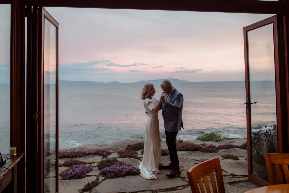 Jemmah and a loved one dancing on the terrace beneath a pink sky, at Thalia Haven on the East Coast of Tasmania, Australia, for an Elopement Collective elopement. Photograph by Joey Willis.