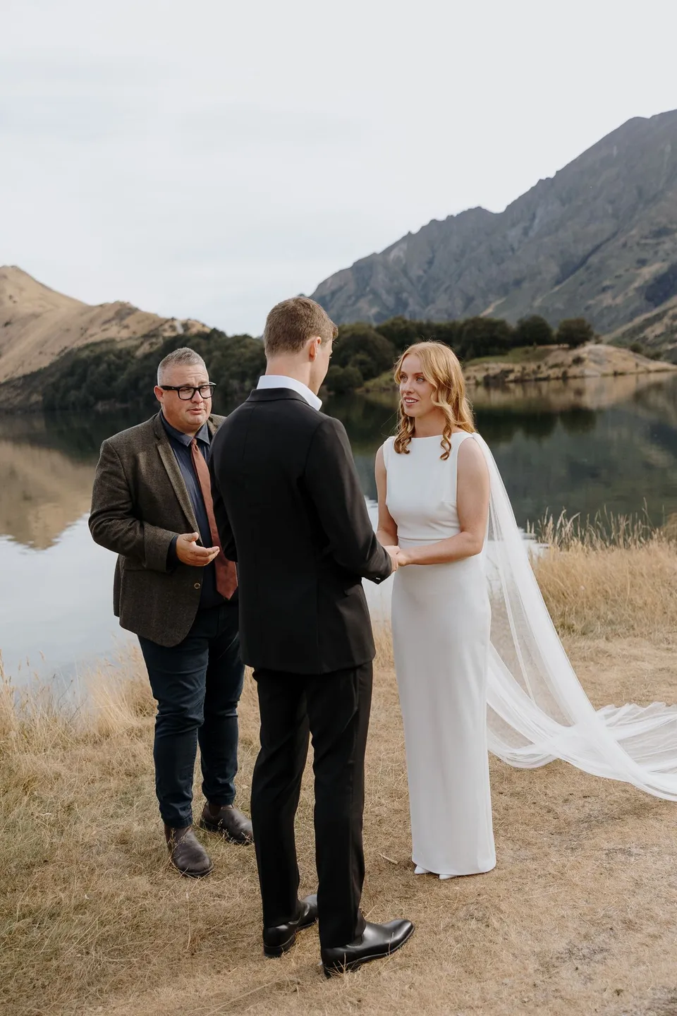 Celebrant Josh Withers leading Neve and Hamish’s lakeside ceremony, with Neve’s white cape trailing behind her, at Moke Lake in Queenstown, New Zealand, for an Elopement Collective Queenstown elopement. Photo by George Bowden.
