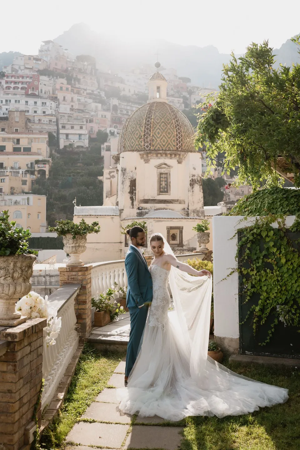 Skyler and Zach standing together beside the terrace balustrade, with her veil lifted to the side and the tiled church dome behind them, at Hotel Buca di Bacco in Positano, Amalfi Coast, Italy, for an Elopement Collective elopement. Photograph by Jason Corroto.