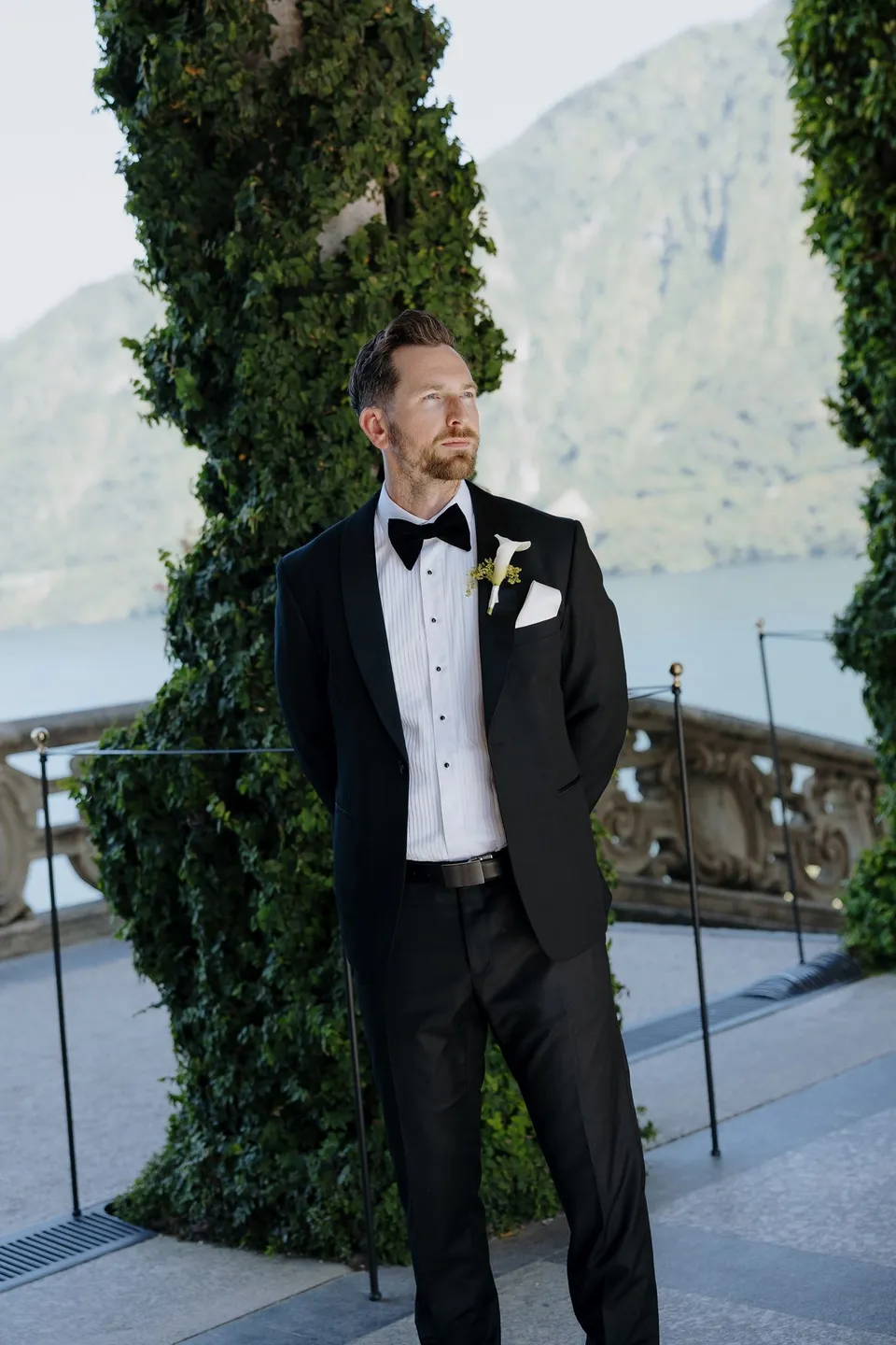 Lachlan in a black tuxedo and black bow tie beneath Villa del Balbianello’s ivy loggia, wearing a white calla lily boutonniere by Garden of Love, at Lake Como in Lombardy, Italy, for an Elopement Collective elopement. Photograph by George Bowden.