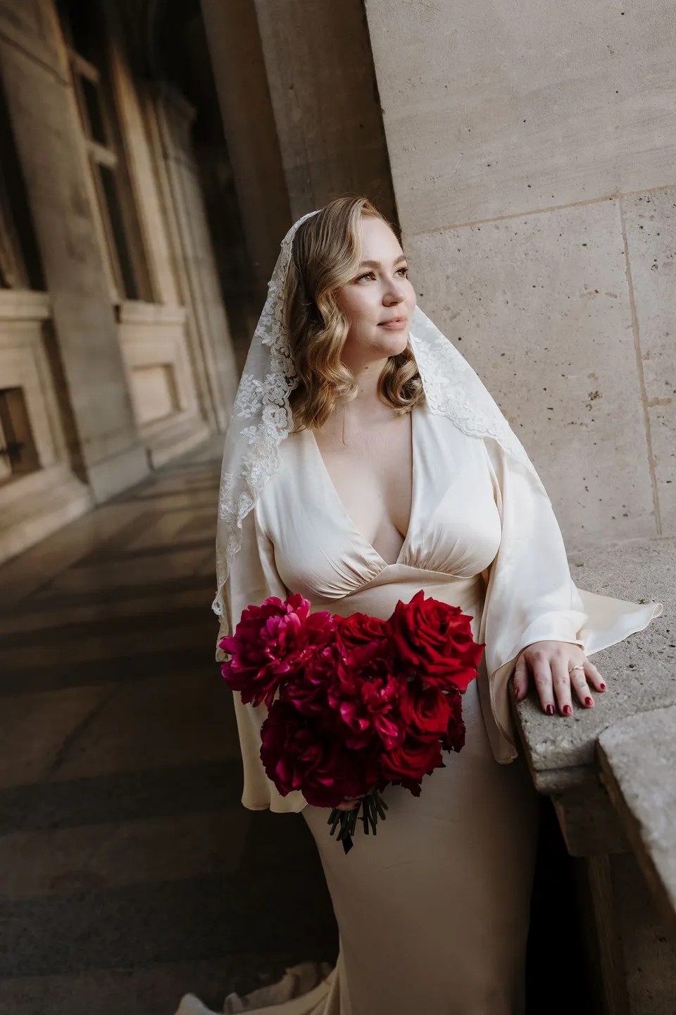 Kara leaning against a stone balustrade beneath the Louvre’s arches, holding her red bouquet by Ivy Bleu, at the Louvre in Paris, France, during an Elopement Collective elopement. Photograph by George Bowden.